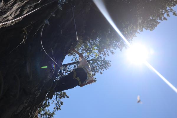 A person is actively climbing a rugged rock face, secured by ropes and carabiners, under a brilliant blue sky